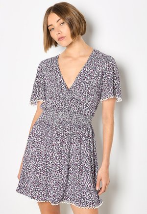 Kobieta ubrana w kwiecistą sukienkę z krótkim rękawem i dekoltem w serek, z talią podkreśloną paskiem oraz falbankami na rękawach i u dołu, stojąca na białym tle.