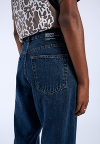 Jeans in denim blu scuro con una finitura texturizzata, dotati di tasche posteriori e di un'etichetta con il marchio in vita. Dettaglio della vista laterale.