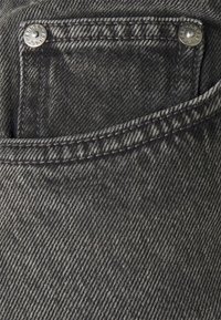 Tmavé džínové kalhoty mají přední kapsu se stříbrným kováním a kontrastním šitím. Textura vykazuje jasný vzor keprové vazby.