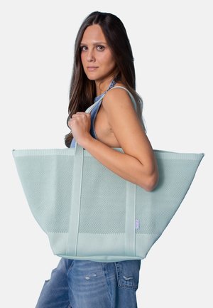 Woman with long brown hair wearing sleeveless top and jeans carries large light blue mesh tote bag over shoulder.