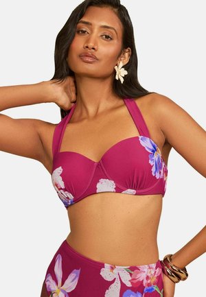 Kobieta w magentowym kwiatowym topie bikini typu halter i majtkach z wysokim stanem, z kolczykami w kształcie kwiatów i złotymi bransoletkami na jednym ramieniu.