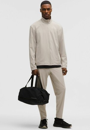 Moški v bež jopi z zadrgo in hlačah, ki drži črno športno torbo, obut v črne superge, stoji pred enobarvnim sivim ozadjem.