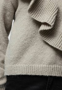 Svijetlosivi pleteni džemper s teksturiranom, uzorkovanom volanom uz jedan bok, s rebrastim rubom i manžetama, upareni s tamnim hlačama.