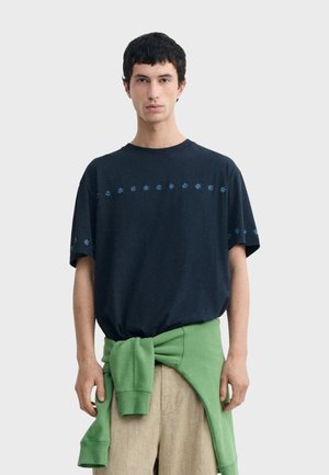 Młody mężczyzna ubrany w granatowy t-shirt z niebieskim wzorem w gwiazdy, beżowe spodnie i zieloną bluzę zawiązaną wokół talii.