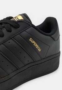 Svart lädersneaker med tre horisontella ränder, guldfärgad "SUPERSTAR"-detalj, texturerad svart gummisula och svarta snören.