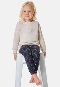 Helles beige Langarmoberteil mit einem kleinen Mondgrafik und dunklen gemusterten Hosen mit Sternen und himmlischen Motiven, an einem barfüßigen Kind.