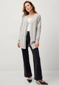 Cardigan aperto grigio chiaro con maniche lunghe e polsini a costine, abbinato a jeans neri a gamba larga e tacchi a punta neri.