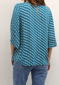 Kvinde iført en teal-bluse med hvidt kattemønster og detalje med nøglehul i ryggen, parret med blå jeans, vendt væk fra kameraet.