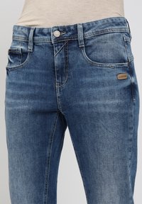 Mittelblaue Jeans aus Denim mit sichtbaren Fronttaschen, Gürtelschlaufen, Knopfverschluss und einem kleinen Markenpatch auf der rechten Seite.