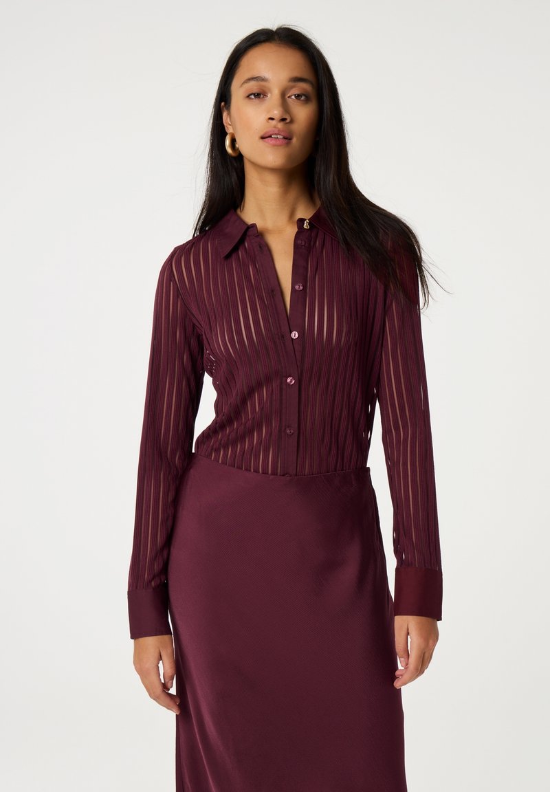 Bordeaux doorzichtige blouse met verticale strepen en lange mouwen, gecombineerd met een bijpassende gladde satijnen rok.
