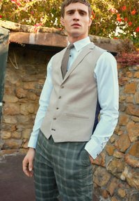 Hellbeige maßgeschneiderte Weste mit vier Knöpfen, kombiniert mit einem hellblauen Hemd und grün karierten Hosen mit einer subtilen Textur.