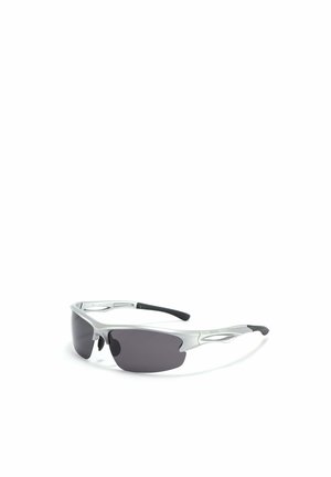 Persona con capelli corti e ricci, che indossa occhiali da sole scuri e un maglione beige a coste con collo, rivolta verso l'obiettivo contro uno sfondo bianco.
