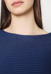 Granatowy prążkowany top z szerokim dekoltem, gładką fakturą i subtelnym połyskiem. Materiał jest miękki i dobrze przylegający, zapewniający komfort.