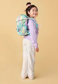 Een jong meisje met een glimlach, gekleed in een pastelpaarse sweater, multicolore gestreepte broek en een grote groene en roze rugzak tegen een beige achtergrond.