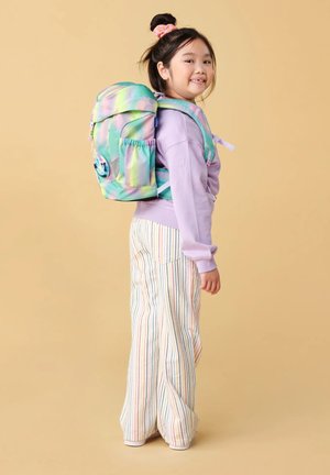Een jong meisje met een glimlach, gekleed in een pastelpaarse sweater, multicolore gestreepte broek en een grote groene en roze rugzak tegen een beige achtergrond.