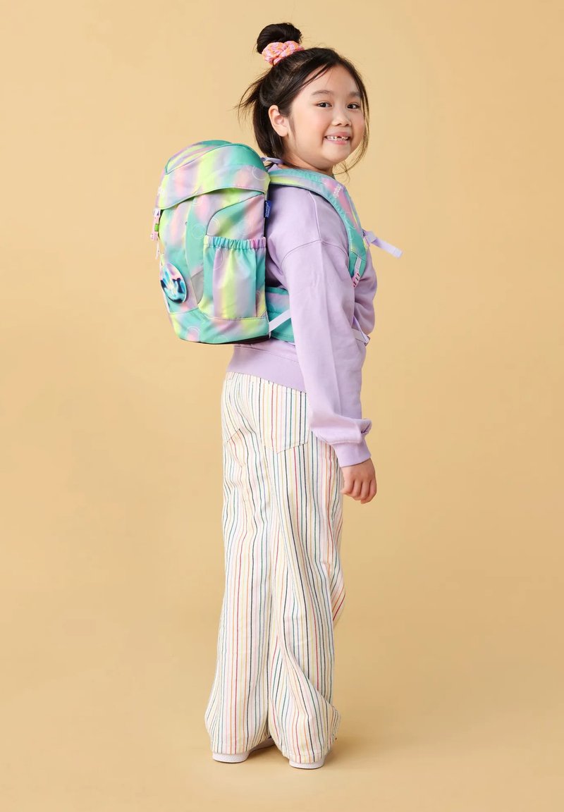 Een jong meisje met een glimlach, gekleed in een pastelpaarse sweater, multicolore gestreepte broek en een grote groene en roze rugzak tegen een beige achtergrond.