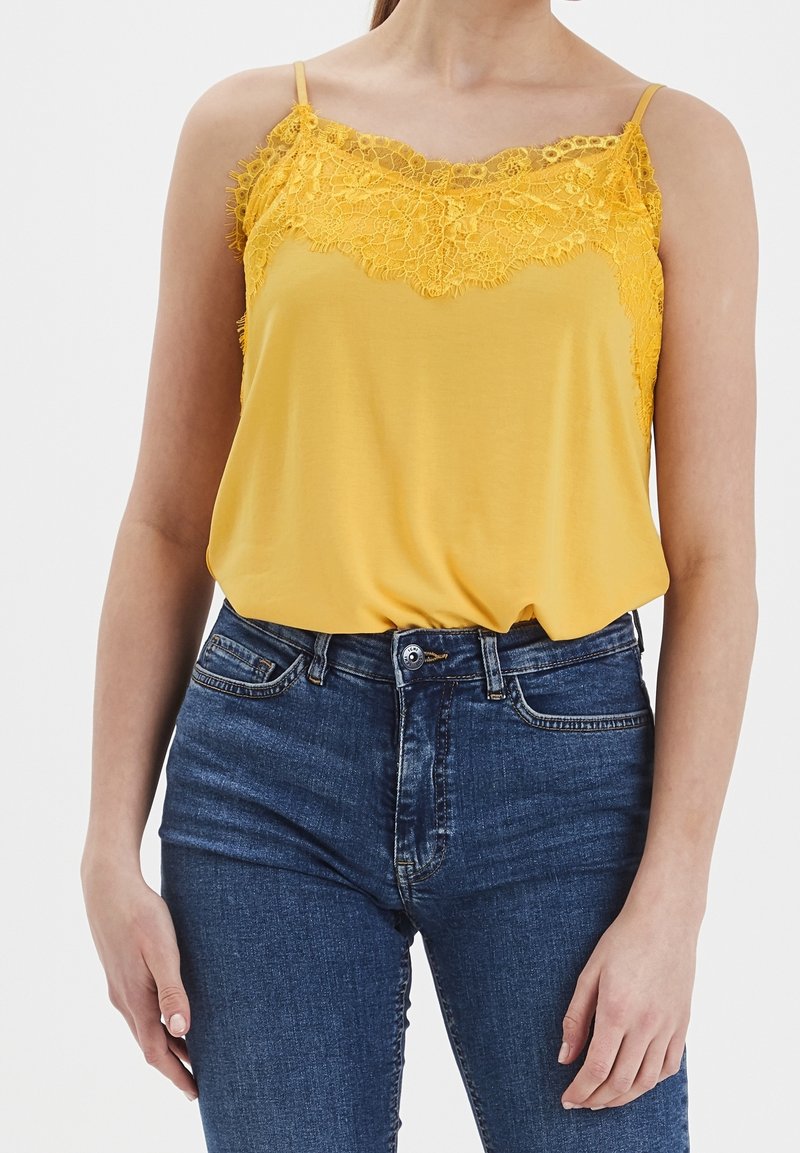 Camisole jaune avec bordure en dentelle au niveau du décolleté, dotée de fines bretelles. Associée à un jean taille haute bleu foncé. Texture de tissu lisse.