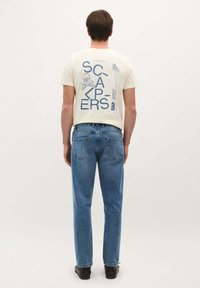 Camiseta de algodón color crema con gráficos azules, que presenta un cráneo y texto, combinada con unos vaqueros de mezclilla azul claro y zapatos negros.