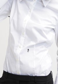 Camisa blanca de botones en tejido ligero, con un diseño de rosa negra bordado en la parte inferior izquierda. Usada con pantalones negros.