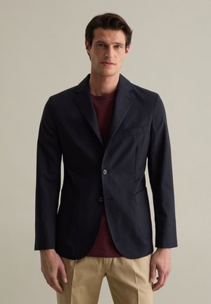 Blazer blu navy con design a una fila di bottoni, lembo a punte, due bottoni e taschino sul petto. Indossato sopra una camicia bordeaux e pantaloni beige.