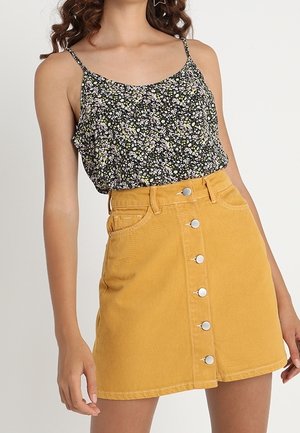 Femme portant un haut sans manches noir à fleurs, rentré dans une mini-jupe jaune moutarde à boutons sur le devant, debout avec un bras détendu.