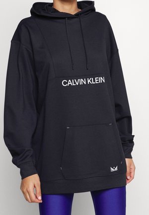 Person iført en sort Calvin Klein hættetrøje med forlomme og hvidt logo, kombineret med skinnende lilla leggings.