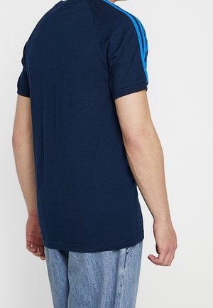 Osoba w granatowym T-shircie z jasnoniebieskimi paskami na rękawach i jasnoniebieskich dżinsach, widziana od tyłu na jednolitym białym tle.