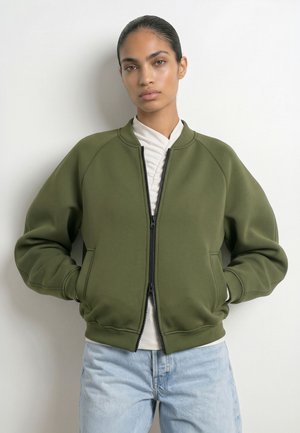 Mladá žena nosiaca olivovozelenú bomber bundu na zips cez biely top a svetlomodré džínsy, stojaca pri obyčajnej bielej stene.