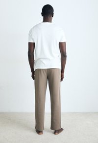 Weißes kurzärmliges T-Shirt kombiniert mit beigen, lässig geschnittenen Hosen; aus weichem Baumwollstoff; mit dem Rücken zur Kamera vor einem hellen Hintergrund.
