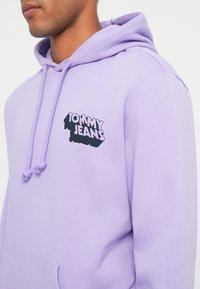 Lila kapucnis pulóver, zsinórral és elülső zsebbel. Fekete "TOMMY JEANS" logó a bal mellrészen. Puha anyag, sima textúrával.
