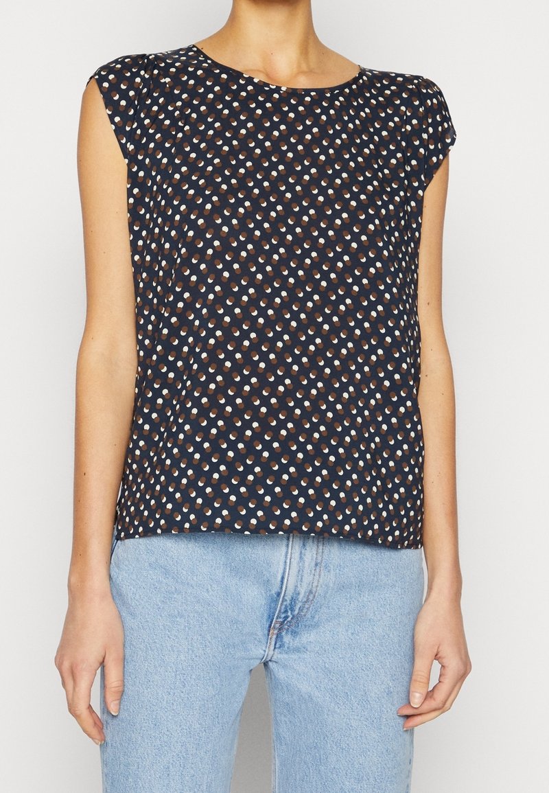 Top sans manches bleu marine avec des pois blancs et marron, encolure ronde, tissu doux, coupe légèrement ample, porté par un modèle.