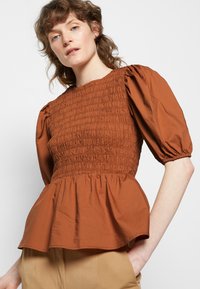 Top de cor ferrugem com corpete franzido e mangas bufantes, finalizado com uma bainha peplum. Feito de tecido texturizado, apresenta um decote redondo.
