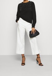 Kvinna som bär en svart tröja med pärldetaljer, vita vida byxor, svarta högklackade sandaler och håller en svart kuvertväska.