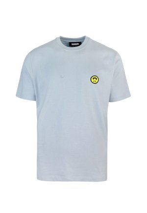 Camisa de algodón de color azul claro con mangas cortas, que presenta un parche circular negro con una cara sonriente amarilla en el lado izquierdo del pecho.