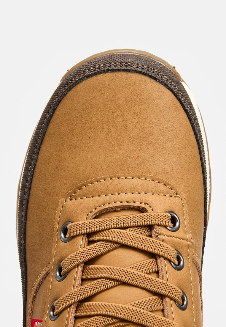 Zapatilla de deporte de imitación de cuero marrón con punta redonda, acentos marrones oscuros, cordones texturizados y ojales circulares. Suela blanca visible en la parte inferior.