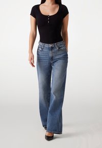 Body contour nero a coste con maniche corte e dettaglio a bottoni, abbinato a jeans a gamba larga blu chiaro. Il modello indossa tacchi neri a punta.