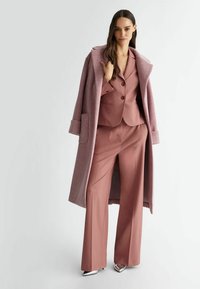 Completo su misura rosa con giacca a un bottone e pantaloni a gamba larga, abbinato a un lungo cappotto rosa chiaro e decolleté a punta argentati.