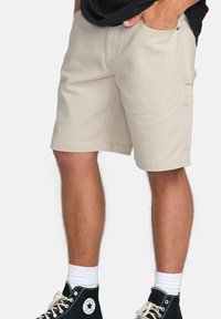 Beige cargo shorts lavet af bomulds-Canvas, med sidelommer og en løs pasform. Vist med sorte højtop sneakers og hvide sokker.