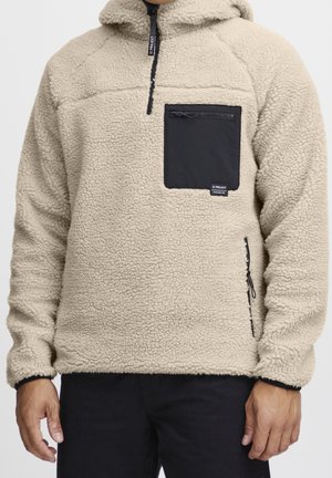 Beige fleece trui met zwart ritssluiting borstzakje en ritssluiting zijzakken, gedragen door een persoon met zichtbare handen.