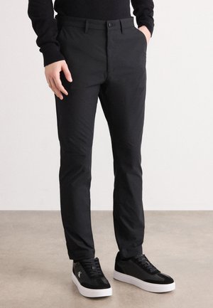 Persoon in slim-fit zwarte broek en zwarte sneakers met witte zolen, staand op een beige vloer tegen een plain witte muur.