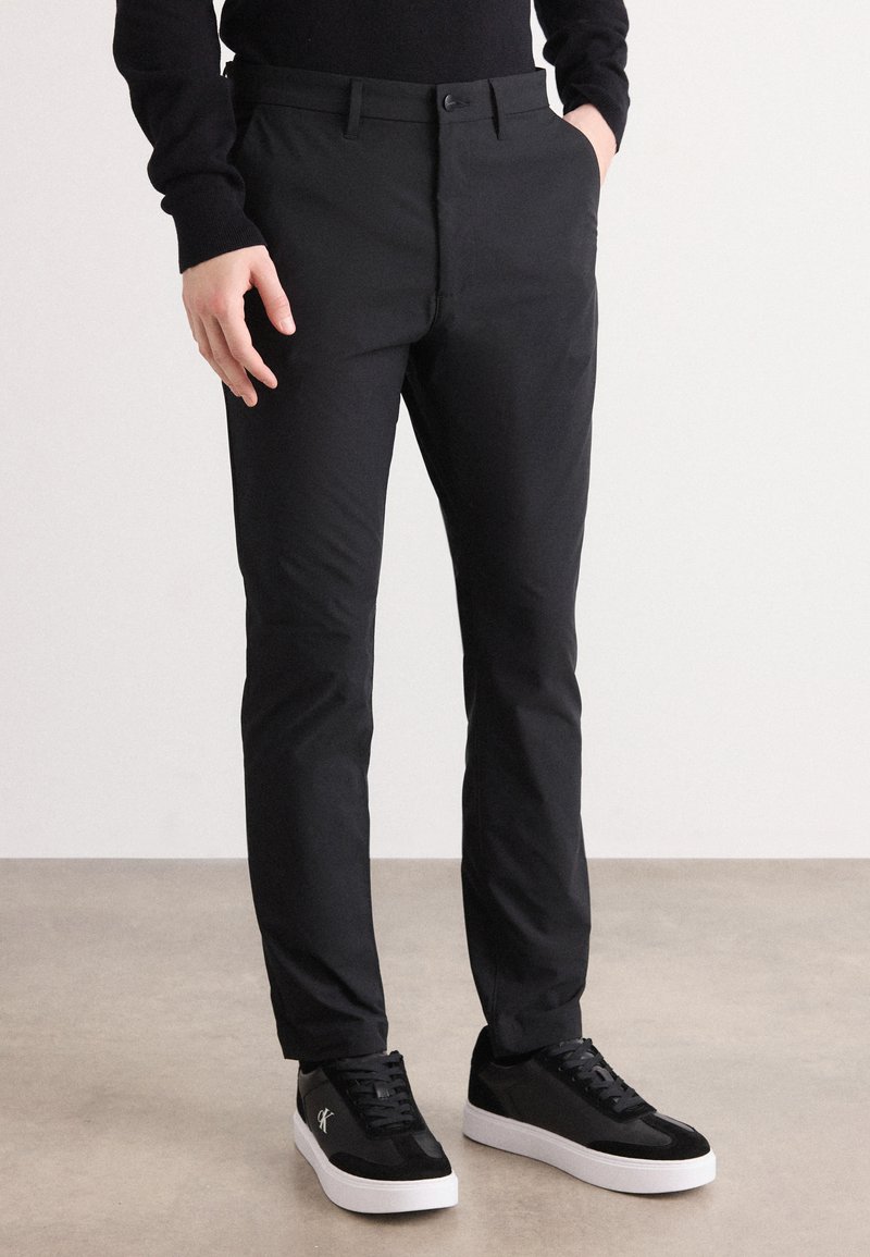 Persona usando pantalones negros ajustados y zapatillas negras con suelas blancas, de pie sobre un suelo beige frente a una pared blanca lisa.
