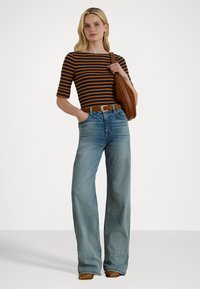 Ženska nosi rjavo in črno prugasto majico, hlače z visokim pasom in širokimi nogami v modri barvi, rjav pas, rjav nahrbtnik čez ramo in rjave čevlje.