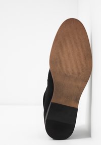 Chaussure en daim noir avec une semelle texturée. La semelle est marron avec des rainures et une finition mate, affichant la taille 42 estampillée dessus.