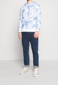 Jasnoniebieski hoodie tie-dye z białymi akcentami, z kapturem na sznurki i nadrukiem z przodu. Noszony z ciemnymi jeansami z mankietami i beżowymi trampkami.
