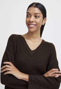 Suéter de color marrón oscuro con cuello en V y mangas anchas, que presenta un patrón de punto texturizado. Diseño simple, sin herrajes visibles.