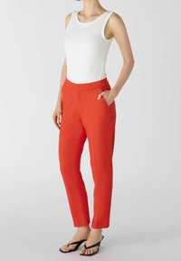 Pantalons rouges ajustés avec poches avant, en tissu mélangé de coton, jambe fuselée, texture lisse, associés à un haut sans manches blanc et des tongs noires.