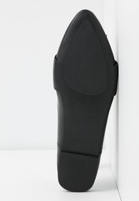 Semelle de chaussure noire à bout pointu avec un talon bloc reposant verticalement contre un angle blanc entre le mur et le sol.