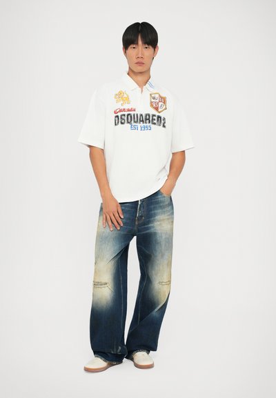 Cămașă polo albă cu logo și insigne, asociată cu blugi din denim deteriorați, de la albastru deschis la albastru închis. Modelul poartă adidași albi.