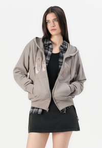 Ung kvinne med langt mørkt hår bruker en beige GAP zip-up hettegenser, svart tanktop, svart-hvitt rutete skjorte og svart skjørt.