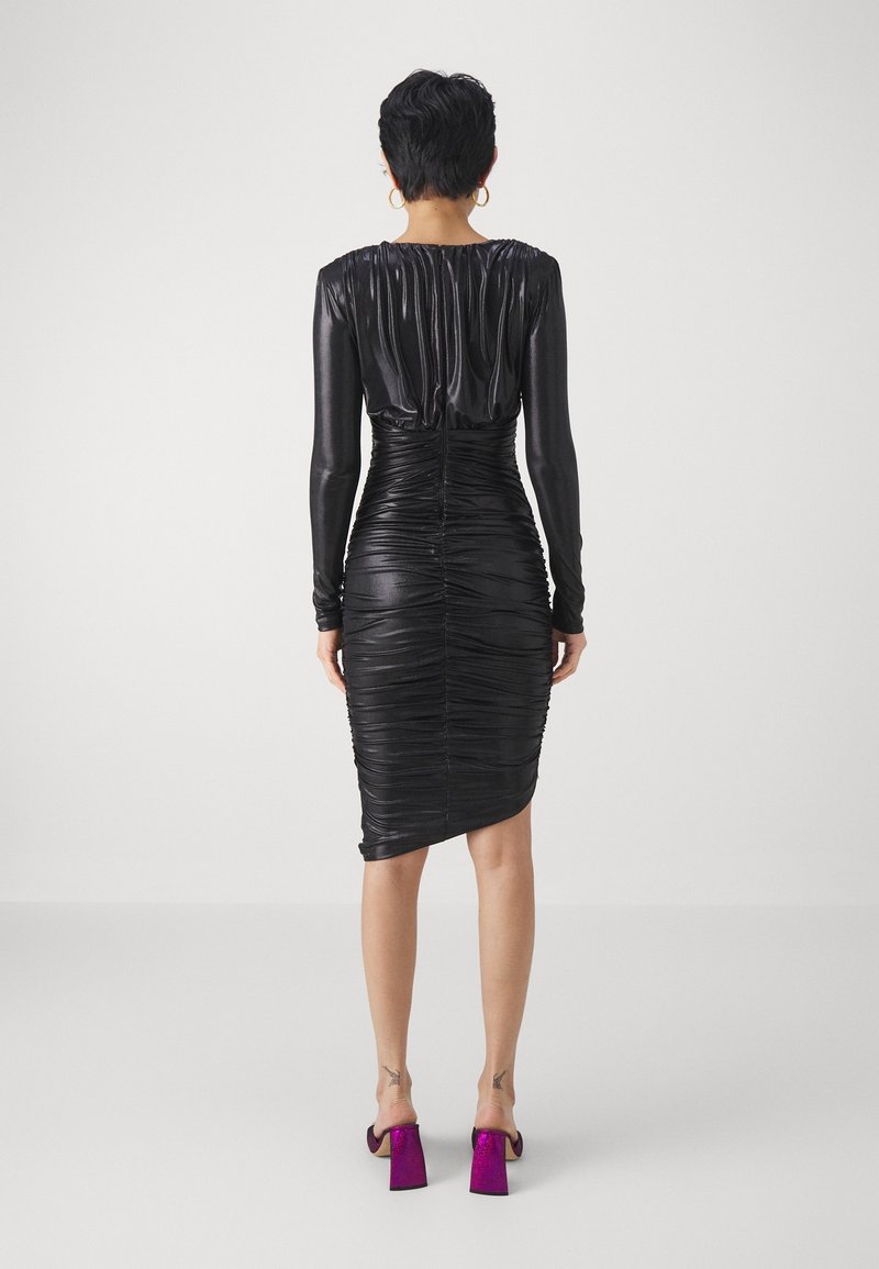 Femme aux cheveux noirs courts portant une robe noire froncée à manches longues et des chaussures à talons blocs violettes, debout de dos sur un fond uni.
